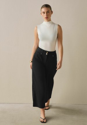 Femeie mergând înainte, purtând un top alb fără mâneci cu gât înalt, pantaloni negri cu talie înaltă și croială largă, sandale negre și o brățară aurie.