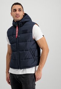 Námornícky modrá puff vestu s kapucňou, so zipsom a bočnými vreckami. Obsahuje látkovú nášivku s logom a červený zips s logom.