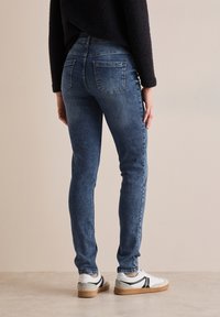 Jeans ajustados de mezclilla azul con bolsillos traseros, cintura de altura media y un ligero estiramiento; combinados con zapatillas blancas con acentos negros.
