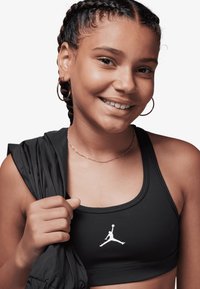 Sutien sport negru cu logo Air Jordan alb, confecționat dintr-un material neted. Modelul are părul împletit și ține o jachetă neagră cu o mână.