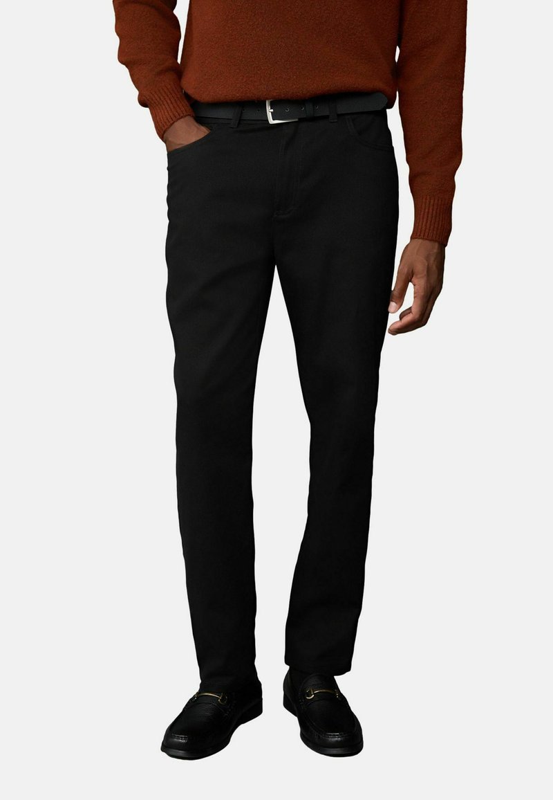 Schwarze Baumwollhosen mit geradem Schnitt, ausgestattet mit seitlichen Taschen und einem Gürtel. Dazu wird ein strukturiertes, rostfarbiges Sweatshirt und schwarze Schuhe getragen.