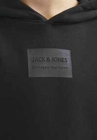 Sudadera negra con un parche de goma texturizado que presenta el logo "JACK & JONES" y "Nuestro Legado Tu Futuro" en letra en relieve.