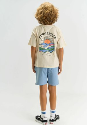 Child with curly hair stands facing away, wearing beige T-shirt with colorful sun and mountains design and words "Surrender and let the sun set in."