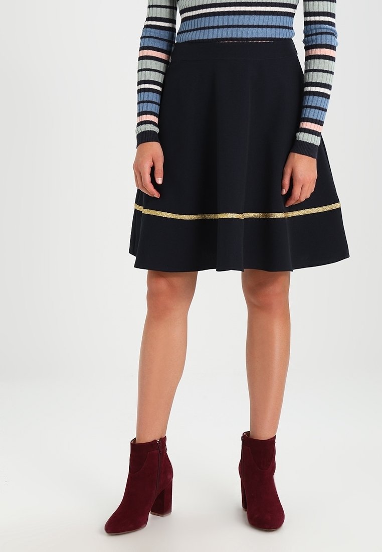 Jupe évasée noire avec une bande horizontale dorée, assortie à des bottines bordeaux. Un haut long à rayures en tricot est visible.