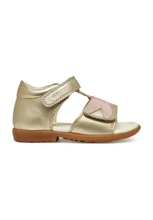 Sandalia de cuero dorado con una tira de velcro, detalle de corazón rosa, diseño de punta abierta y suela de goma para agarre y comodidad.