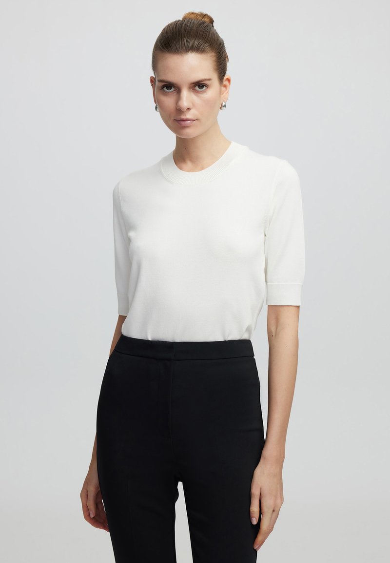 Top de punto blanco de manga corta con cuello redondo, combinado con pantalones negros de talle alto y corte entallado. Textura suave y silueta ajustada.
