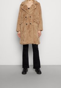 Brązowy dwurzędowy płaszcz z imitacji futra z szerokimi klapami, dwiema przednimi kieszeniami i czarnymi guzikami, noszony na czarnych rozkloszowanych spodniach i masywnych butach.