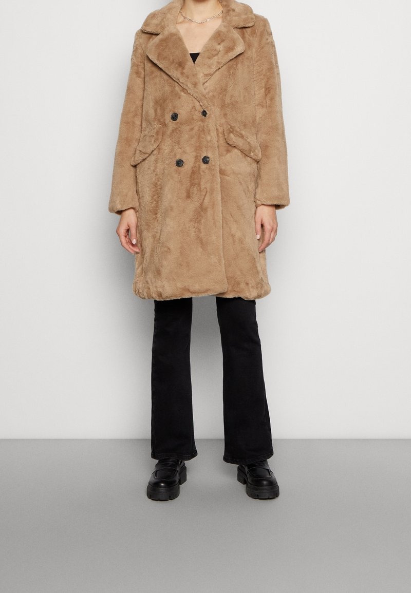 Brązowy dwurzędowy płaszcz z imitacji futra z szerokimi klapami, dwiema przednimi kieszeniami i czarnymi guzikami, noszony na czarnych rozkloszowanych spodniach i masywnych butach.