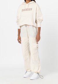 Krämfärgad huvtröja med texten "DICKIES", tillsammans med ljuskrämfärgade cargobyxor med fickor. Vita sneakers kompletterar outfiten.