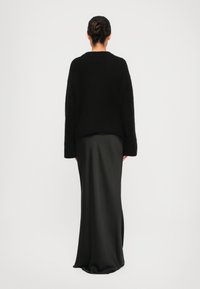 Donna con i capelli raccolti in un chignon, che indossa un maglione nero di pelliccia e una lunga gonna di raso nera, in piedi di spalle contro uno sfondo neutro.