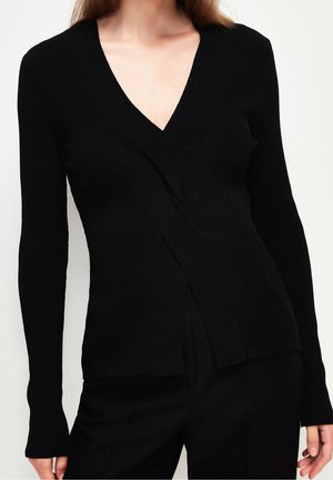 Femme portant un pull en maille côtelée noire à manches longues et col en V, associé à un pantalon noir.