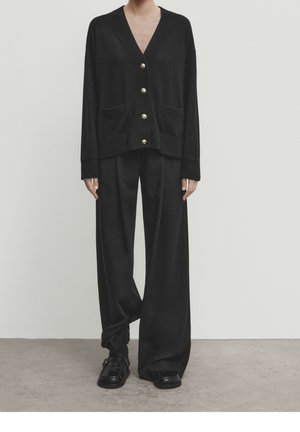 Persoon draagt een losse zwarte vest met knopen en zakken aan de voorkant, bijpassende wijd uitlopende broek en zwarte loafers, staand tegen een effen achtergrond.