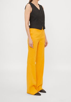 Mujer con chaleco negro sin mangas abotonado y pantalones de pierna ancha de talle alto color mostaza con zapatos negros puntiagudos.