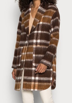 Woman wearing a brown, white, and beige plaid wool coat with lapels, front pockets, and a mid-thigh length over light-colored pants.