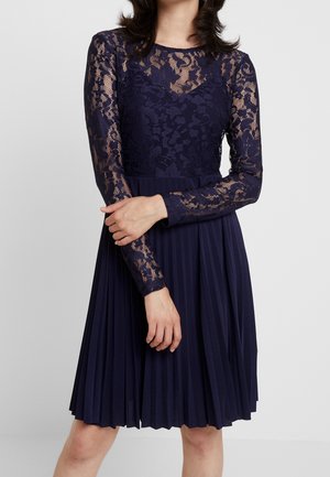 Kvinde iført en knælængde marineblå kjole med lange blonder ærmer og en plisseret nederdel, stående mod en ensfarvet baggrund.