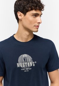 Tengerészkék pamut póló, kerek mintával fényes ezüst színben, rajta a "MUSTANG JEANS COMPANY" felirattal és a megalapítás dátumával.