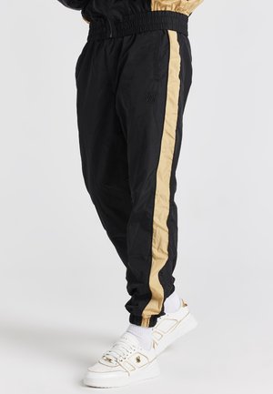 Czarne spodnie dresowe z beżowymi paskami po bokach, noszone z białymi trampkami i białymi skarpetkami na osobie stojącej na jednolitym tle.