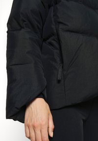 Gros plan sur la main d'une personne à côté du poignet d'une doudoune noire avec une poche zippée, au-dessus d'un pantalon noir.