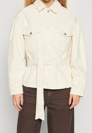 Veste crème boutonnée avec poches à rabat à l'avant et ceinture assortie portée avec un pantalon marron foncé.