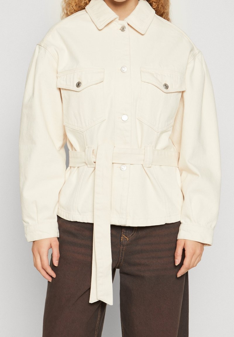 Veste crème boutonnée avec poches à rabat à l'avant et ceinture assortie portée avec un pantalon marron foncé.