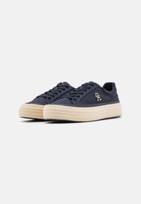 Navy canvas sneakers with natural woven rope sole, flat laces, and a small metallic logo on the side. Rounded toe and low-profile design.