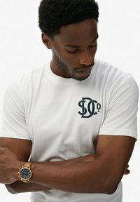 Hvid bomulds t-shirt med et mørkeblåt logo på brystet. Korte ærmer og rund halsudskæring. Båret med et guld ur.
