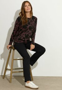 Pull floral bourgogne et noir, pantalon noir et baskets blanches. Le modèle est assis sur un tabouret en bois devant un fond neutre.