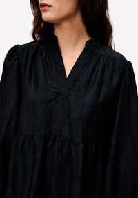 Femme portant un chemisier noir texturé à manches longues avec un col en V, vu des épaules à la taille sur un fond blanc.