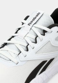 Baskets blanches avec une tige en mesh, accents noirs et lacets. Présente une surface texturée et un logo bien en évidence sur la languette.