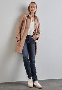 Manteau beige avec des boutons noirs, porté sur un pull léger et un jean foncé. Les chaussures comprennent des baskets montantes blanches et beiges.