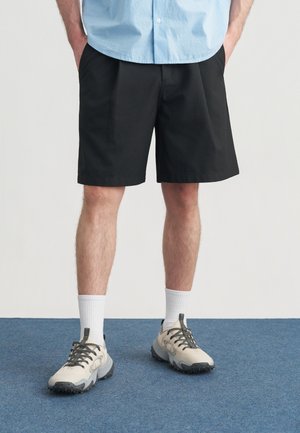 Czarne szorty o luźnym kroju i gładkim przodzie. Noszone z jasnoszarymi sneakersami i białymi skarpetkami typu crew. Tło stanowi niebieski dywan.