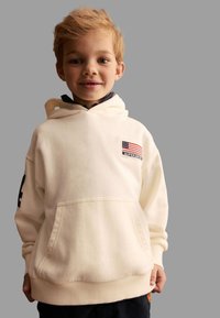 Krämfärgad hoodie med en strukturerad yta, framficka och ett amerikanskt flaggmärke. Har ribbade ärmslut och nederkant.
