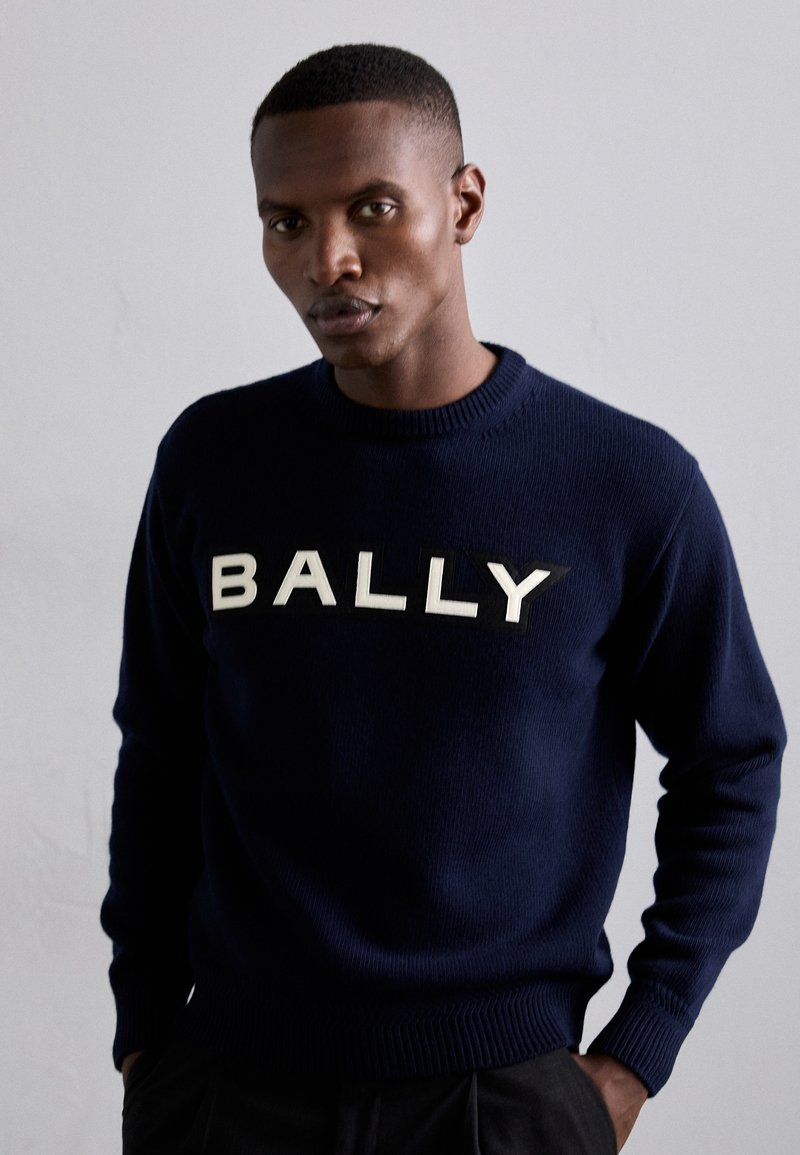 Navy blue knit sweater with ribbed cuffs and hem, featuring bold cream "BALLY" lettering across the chest in a contrasting texture.