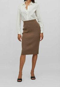 Stickad brun blyertskjol med ribbat mönster, kombinerad med en vit skjorta med knappar. Modellen bär svarta sandaler med öppen tå. Enkel, figurnära design.