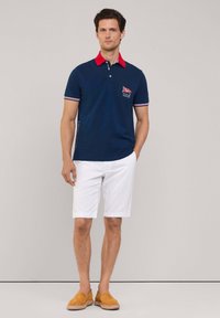 Marineblaues Poloshirt mit rotem Kragen, gestreiften Ärmeln und Brustpatch; kombiniert mit weißen Shorts und hellbraunen Slipperschuhen. Glatte Baumwollstruktur.