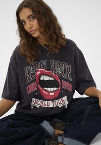 Fiatal nő göndör hajjal, fekete, oversized pólót visel, piros ajkakkal és a "Rebel Rock Tour 1990 World Tour" felirattal, keresztbe tett lábbal ül.