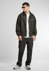 Jachetă neagră ușoară cu închidere prin capse și două buzunare, purtată peste un tricou grafic de culoare gri deschis și pantaloni întunecați, lejeri.