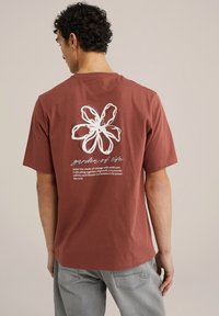 Rozsdabarna pamut póló, fehér virággrafikával és felirattal a hátsó részén. Rövid ujjú, kerek nyakkivágású, laza fit, sima textúrájú.