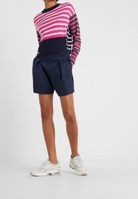 Jambes d'une personne portant un short plissé bleu marine, des baskets blanches épaisses et un pull à manches longues rayé rose, bleu marine et blanc.