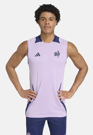 Giovane uomo in piedi con le mani sui fianchi, indossa una canotta sportiva lavanda e pantaloncini blu navy con loghi di gallo sul petto e sulla coscia.