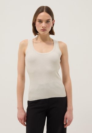 Donna con capelli corti castani, che indossa orecchini a cerchio dorati, un top senza maniche in maglia color crema e pantaloni neri, in piedi davanti a uno sfondo chiaro e uniforme.