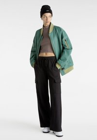 Giacca bomber verde con rifiniture beige, crop top marrone, pantaloni cargo neri e un berretto nero. Caratteristiche: vestibilità rilassata e tasche laterali.
