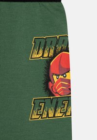 Grüne Baumwollshorts mit einem grafischen Druck eines roten Charakters mit Maske und gelben Augen, sowie dem Text "DRAGON ENEMY" in fettgedrucktem Gelb.