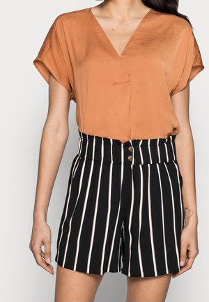 Frau trägt ein lockeres oranges V-Ausschnitt-Top, das in hoch taillierte schwarze Shorts mit weißen vertikalen Streifen und Knöpfen gesteckt ist.