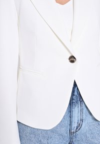 Blazer blanc avec un bouton argenté, épaules structurées et deux poches à l'avant, associé à un jean en denim bleu clair.