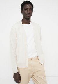 Lys beige kardigan med v-hals, knappfront og ribbestrikkede mansjetter. Brukt over en hvit t-skjorte og sammen med lys beige bukser.
