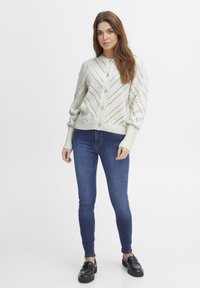 Cardigan tricoté de couleur claire avec des poignets côtelés et une fermeture par boutons, présentant un motif en chevrons. Porté par-dessus un jean skinny en denim foncé.