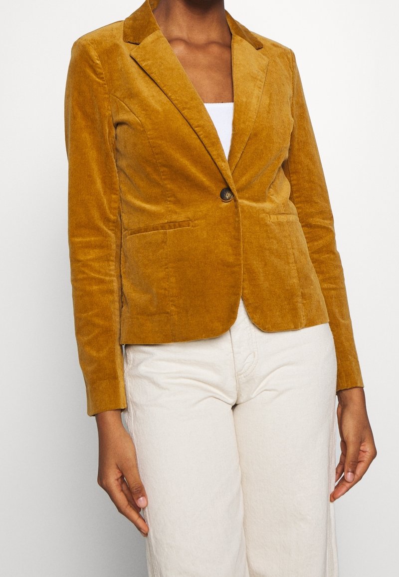 Femme portant un blazer en velours jaune moutarde avec un bouton et un pantalon blanc, debout devant un fond uni.