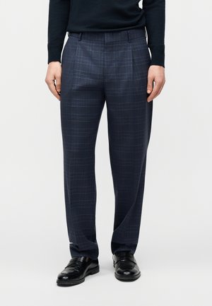 Mężczyzna w granatowych, kraciastych spodniach, czarnych mokasynach i czarnym długim rękawie stoi na jednolitym tle.