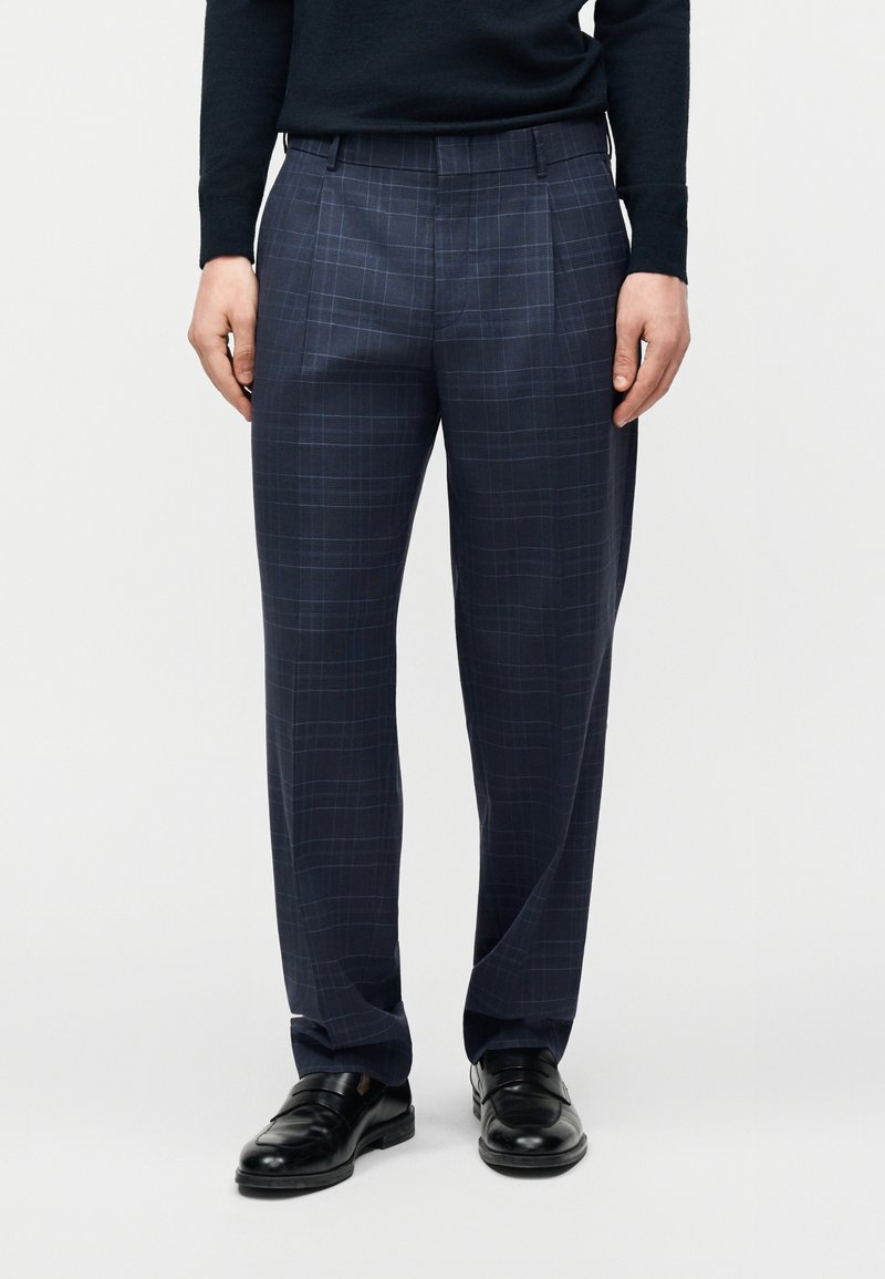 Άνδρας που φοράει καρό παντελόνια σε ναυτικό μπλε, μαύρα μοκασίνια και μαύρο μακρυμάνικο τοπ, στέκεται μπροστά σε ένα απλό φόντο.